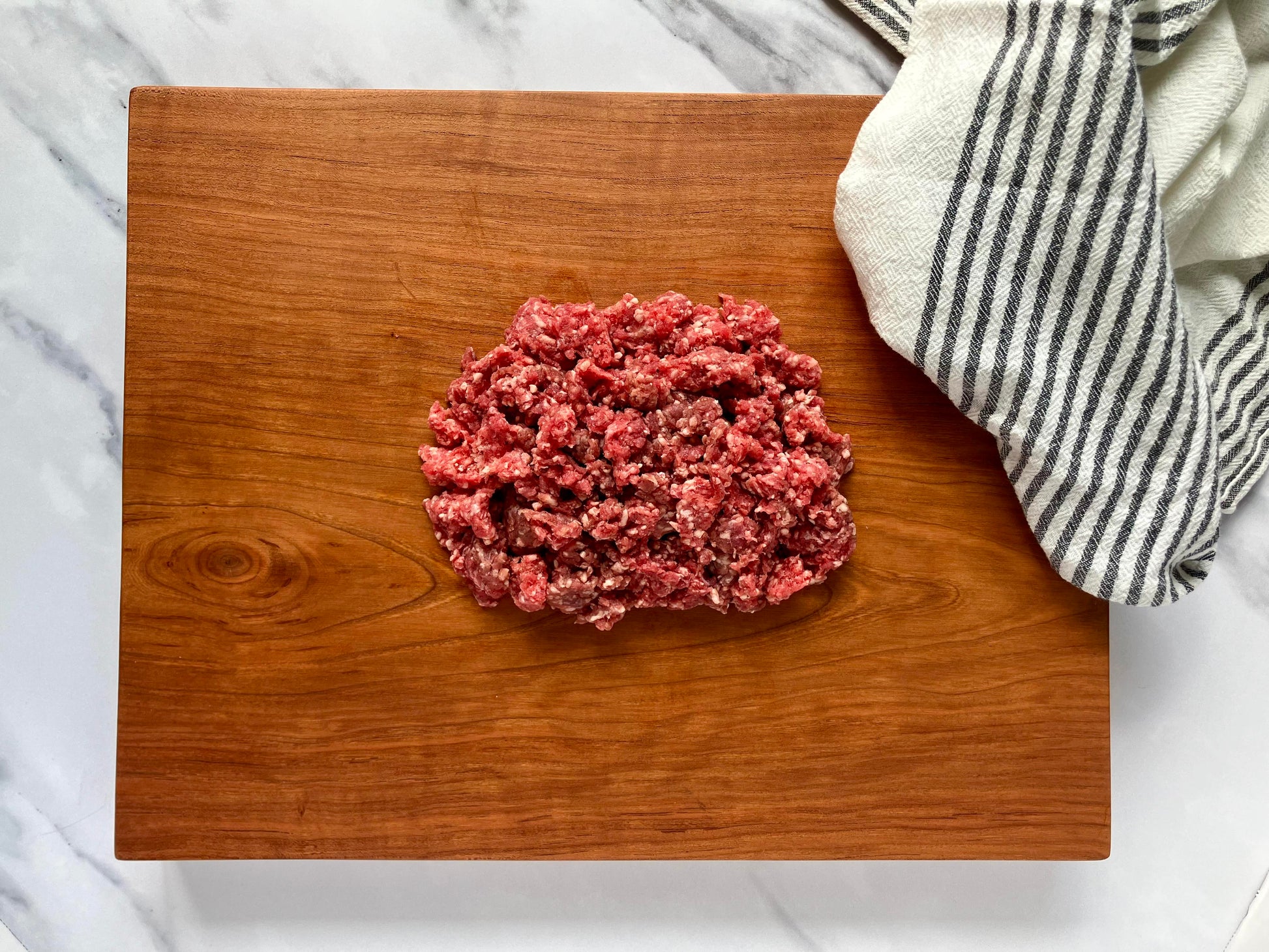 Raw grass fed ground beef on a wooden cutting board with a striped towel in the background.